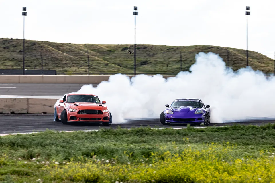 Stefan's Mustang drifting in tandem with a purple Corvette, both cars trailing tire smoke